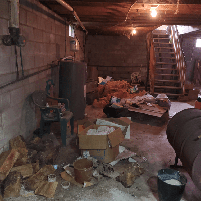 Cluttered basement with exposed pipes, wood logs, and scattered debris. Image is rotated